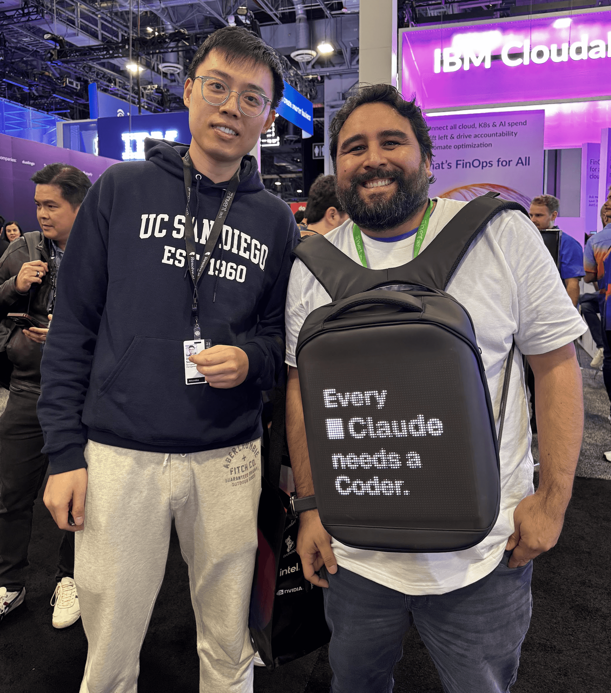 2 people in a packed conference hall. The person on the right is wearing a backpack on their chest with a digital display saying 'Every Claude needs a Coder.'