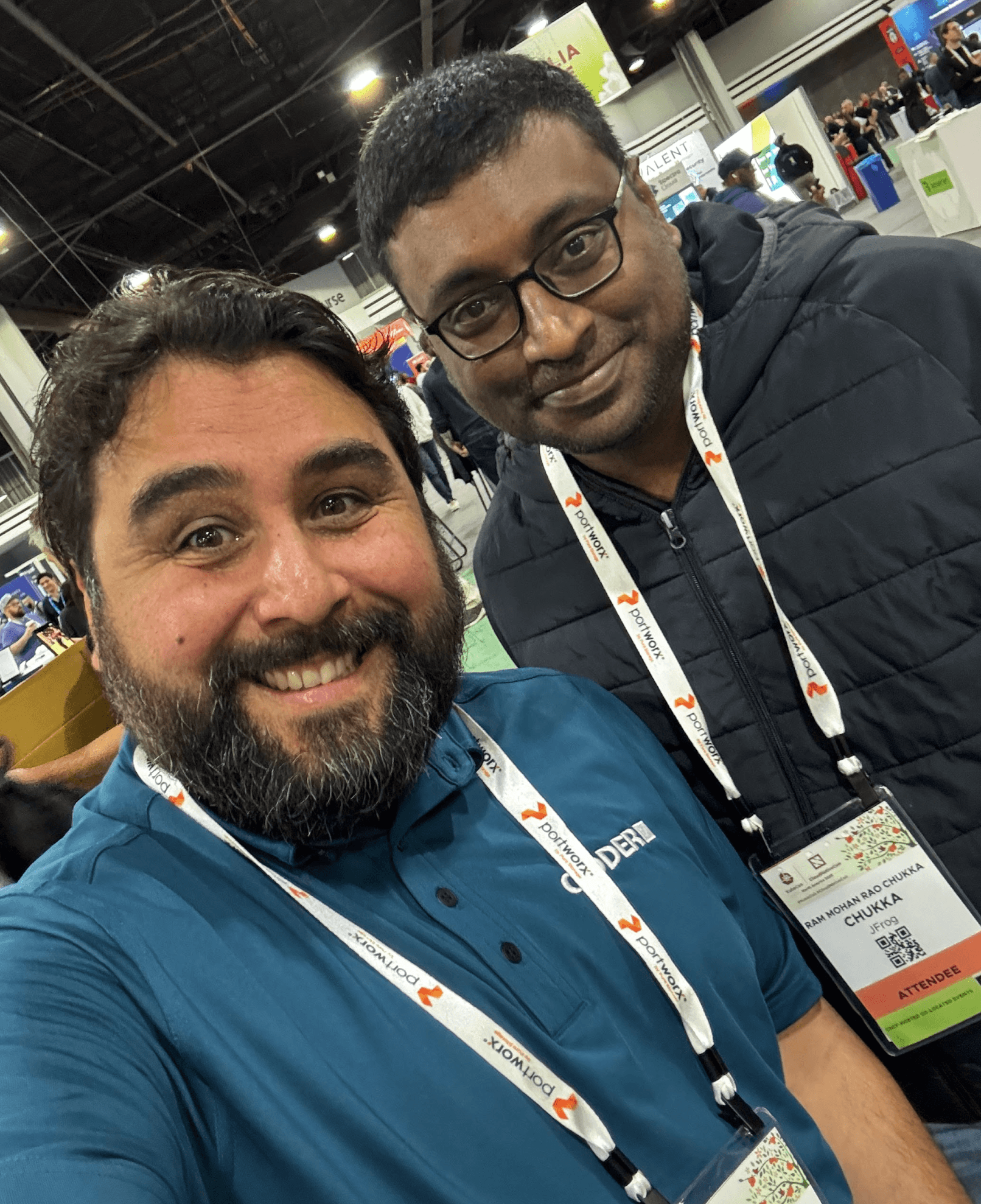 2 people in lanyards smiling together for a selfie in a conference hall. The person on the left is wearing a Coder shirt.