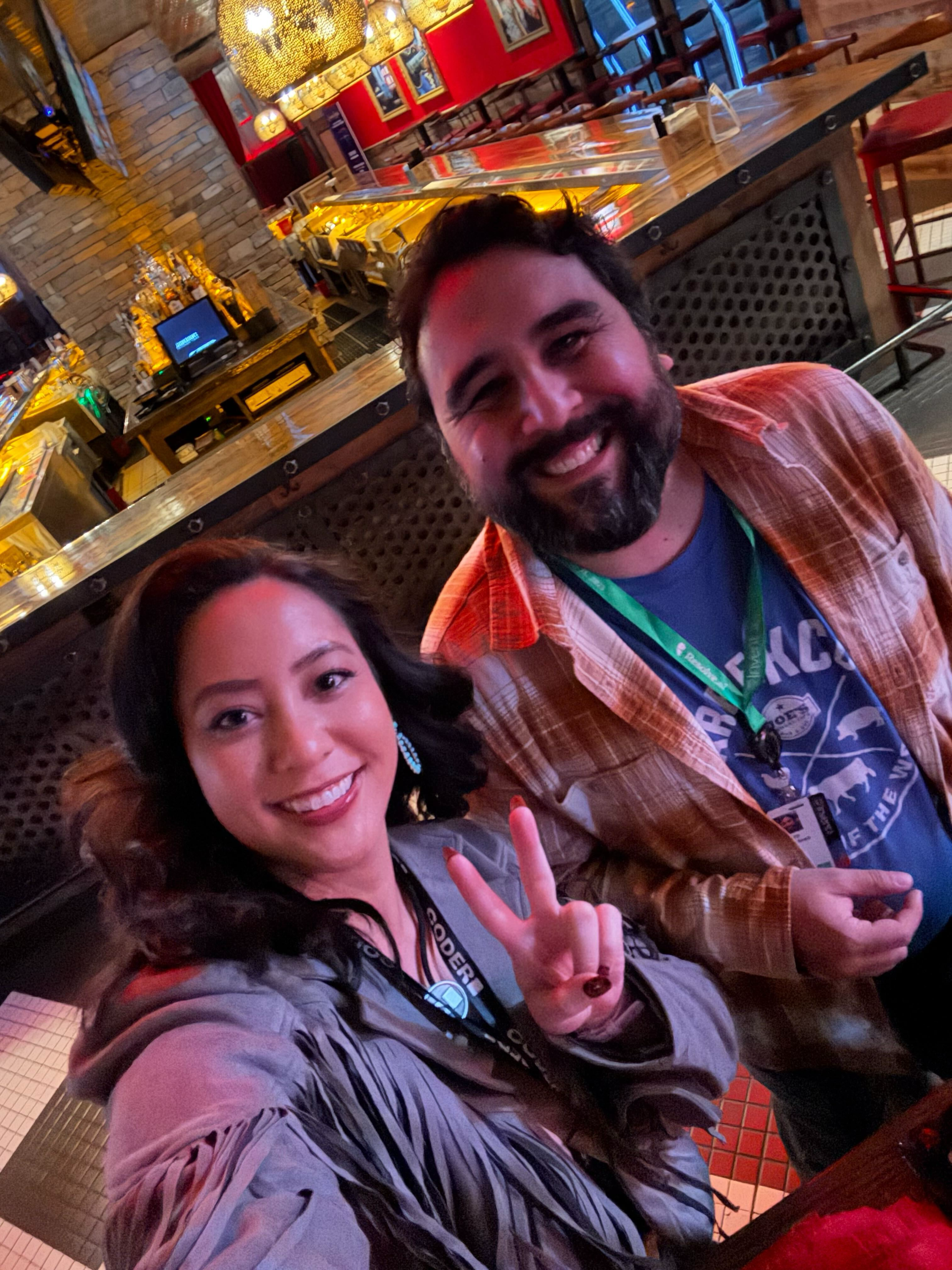 2 people in lanyards smiling in a selfie in front of a bar