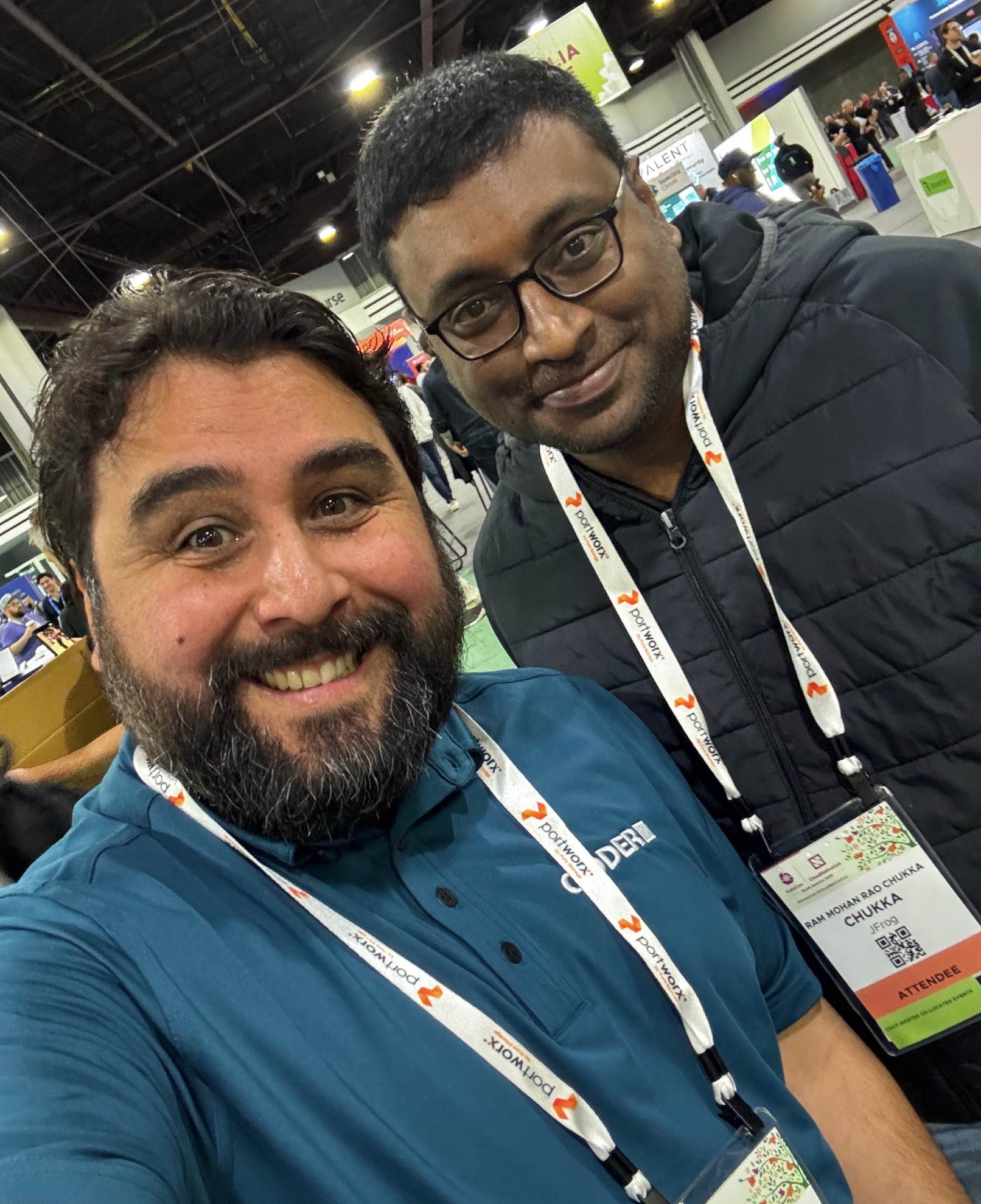 2 people in lanyards smiling together for a selfie in a conference hall. The person on the left is wearing a Coder shirt.