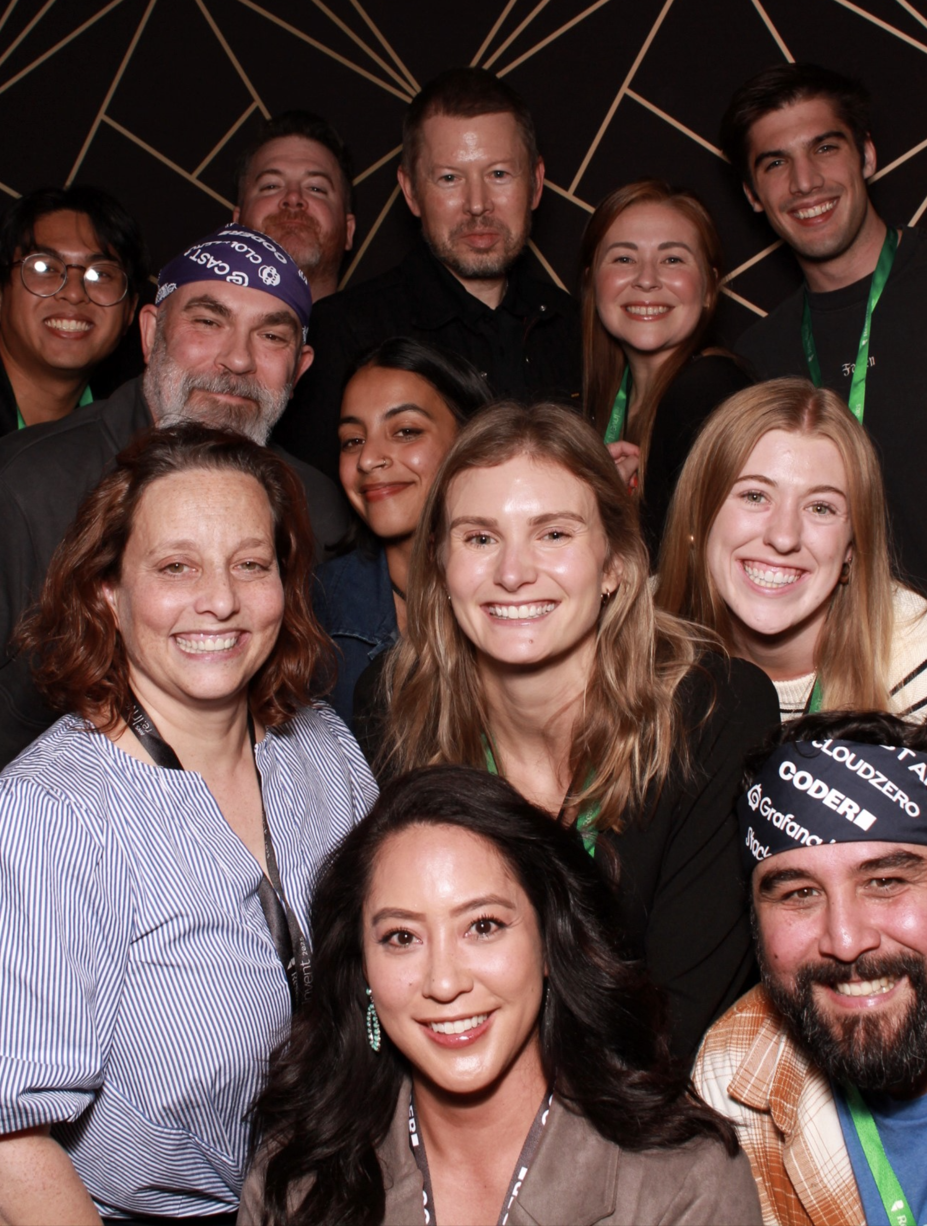 group of 12 people smiling together. 2 are wearing headbands with company logos including Coder, CloudZero, and Grafana