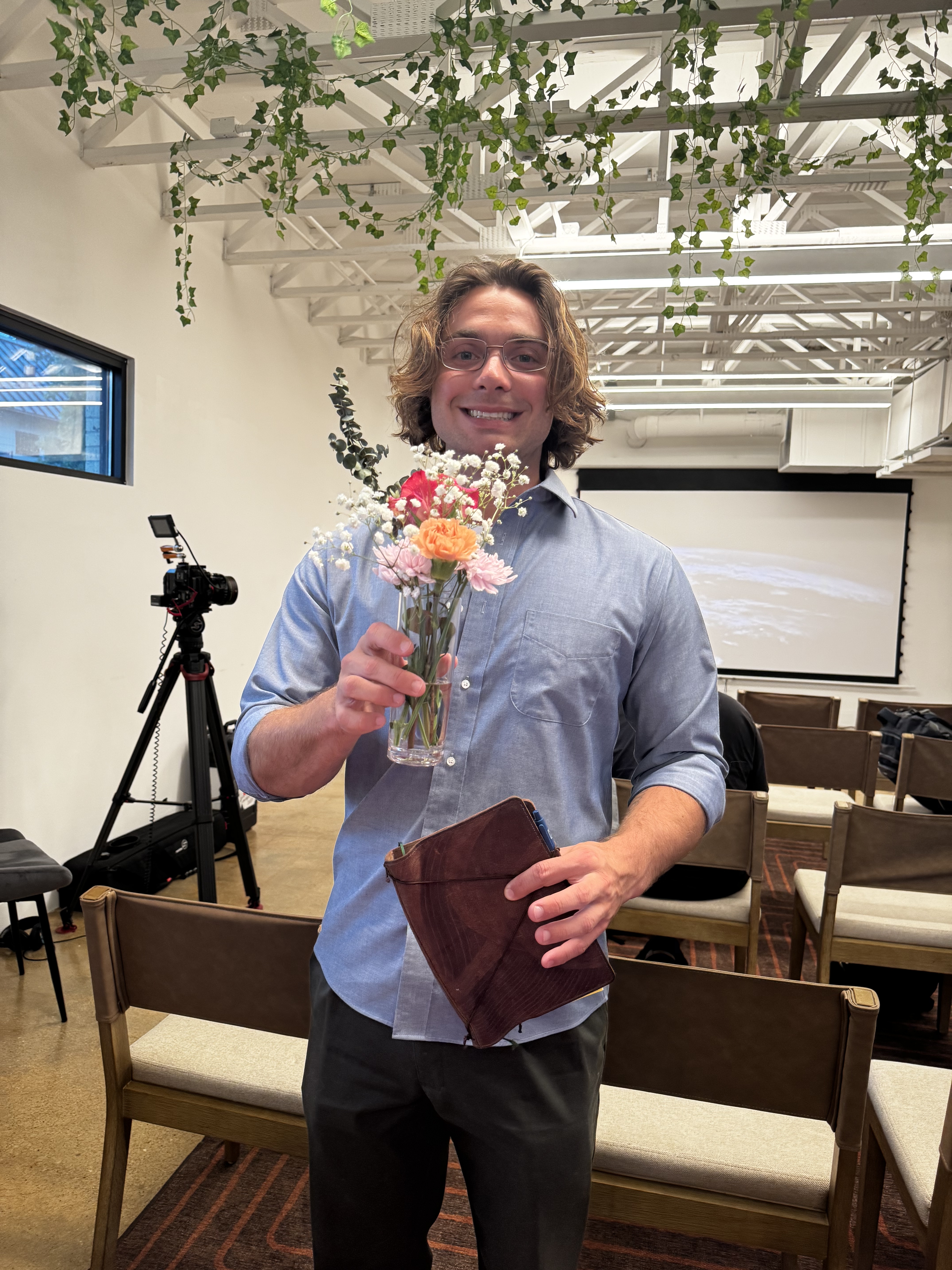 person smiling while holding a notebook in one hand and a small vase of flowers in the other hand